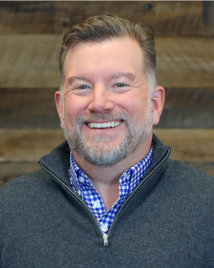 Mike Flanagan, a white man in a gray sweater, smiles. He stands against a shiplap wood background.